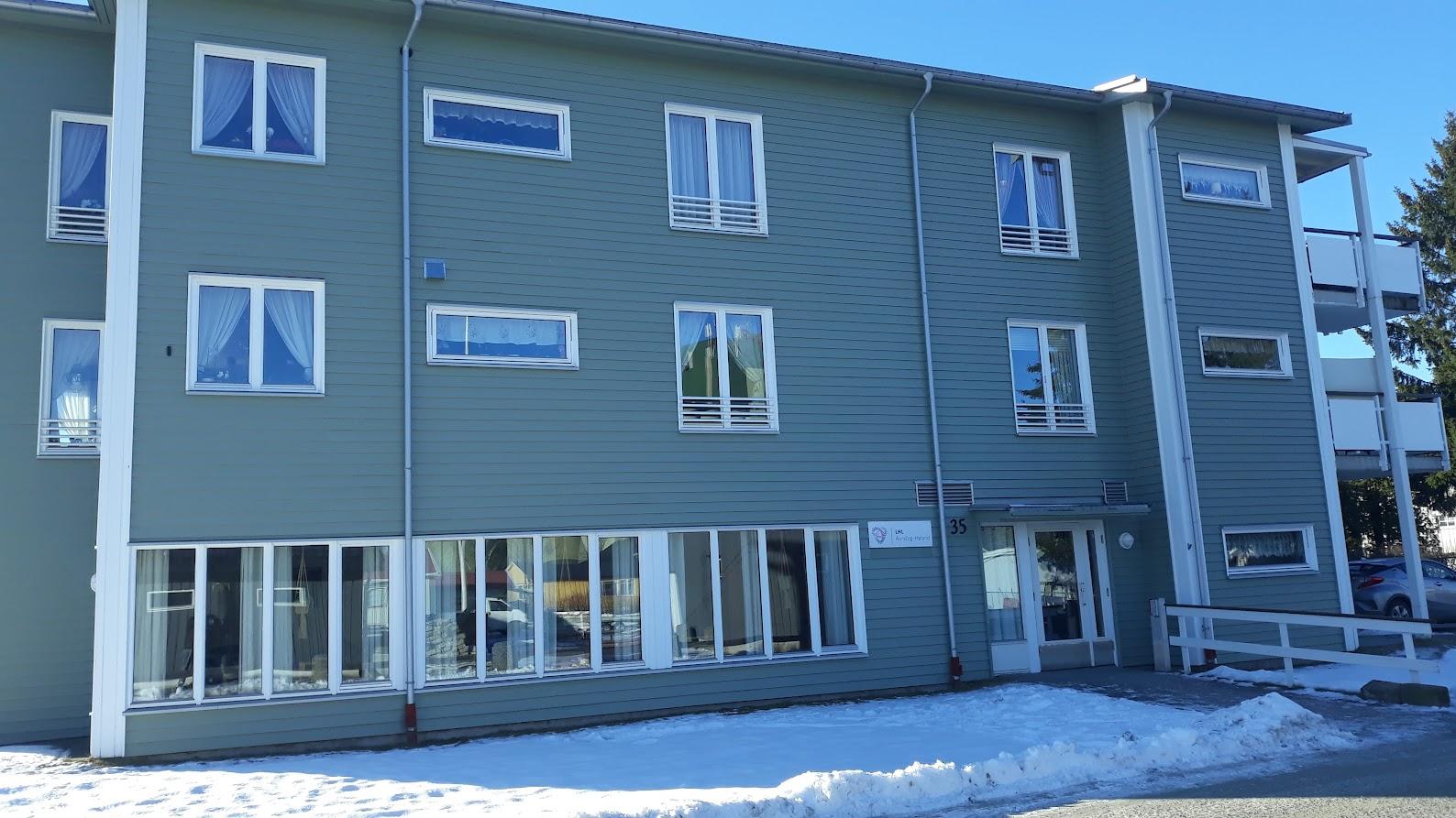 Moderne leilighetsbygg med grønne vegger og snø foran, beliggende i et solfylt område.