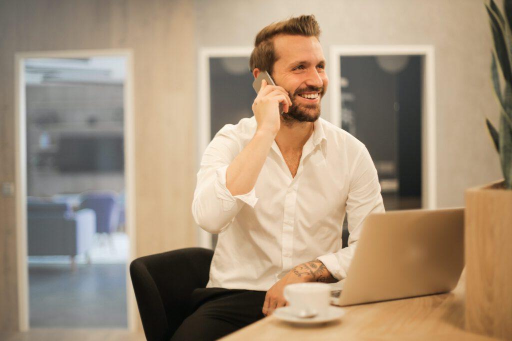 En smilende mann i hvit skjorte snakker i telefonen mens han sitter ved en laptop med en kaffekopp ved siden av seg.