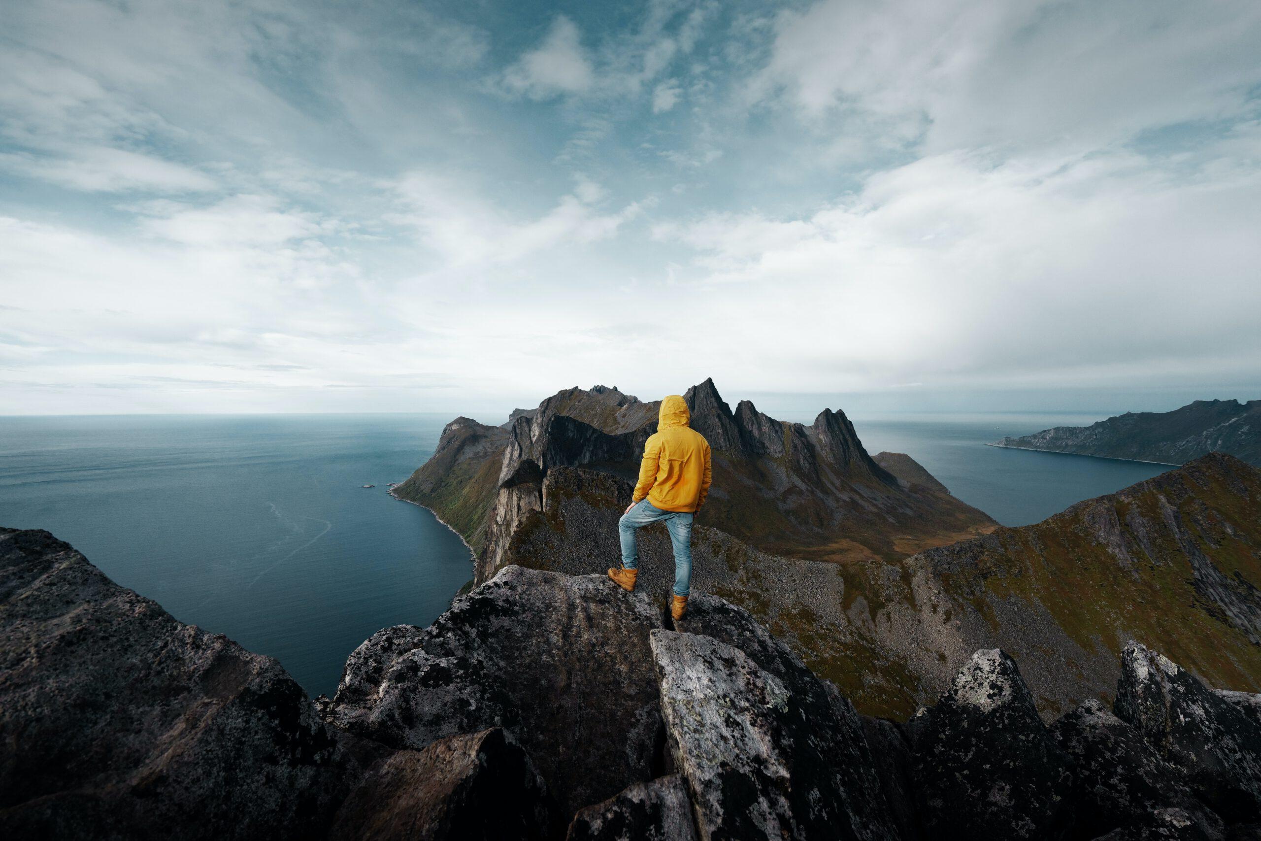 En person i gul jakke står på en fjelltopp med utsikt over havet og fjellene i bakgrunnen.