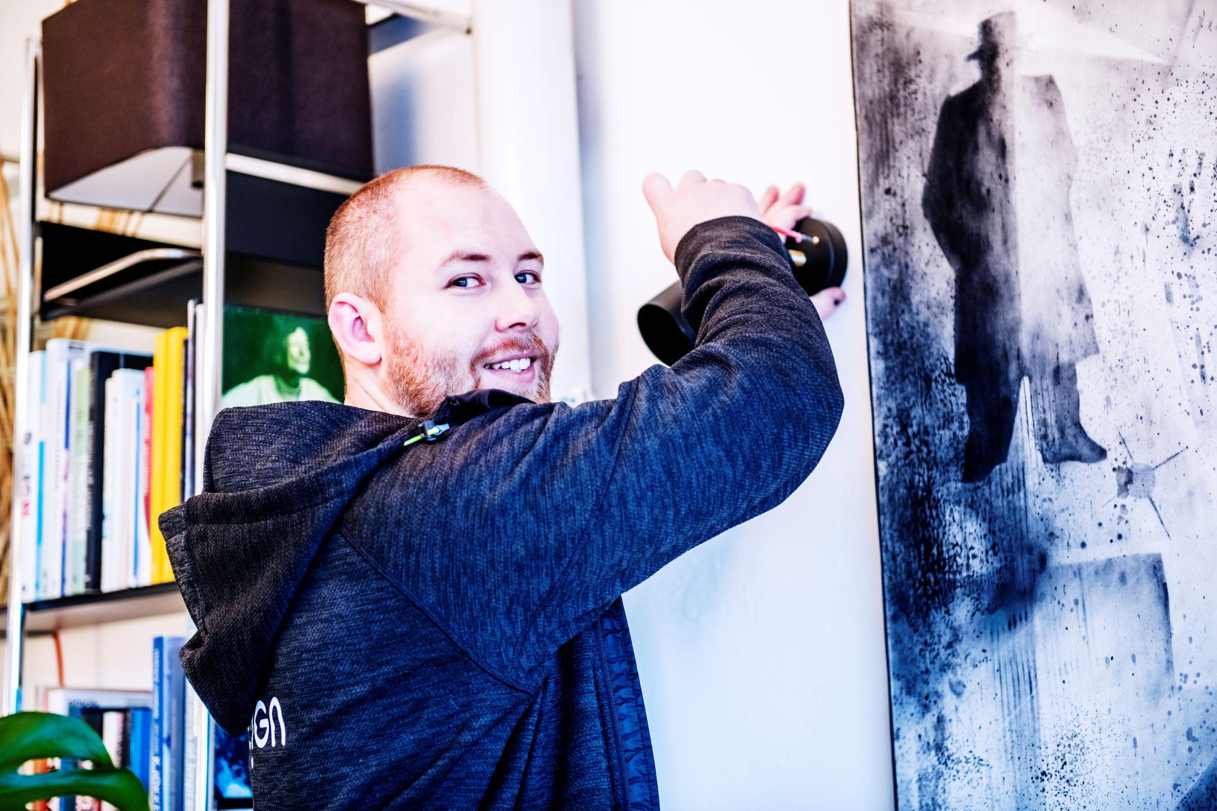 En smilende mann henger opp en ramme på veggen i et rom med bøker og kunstverk.