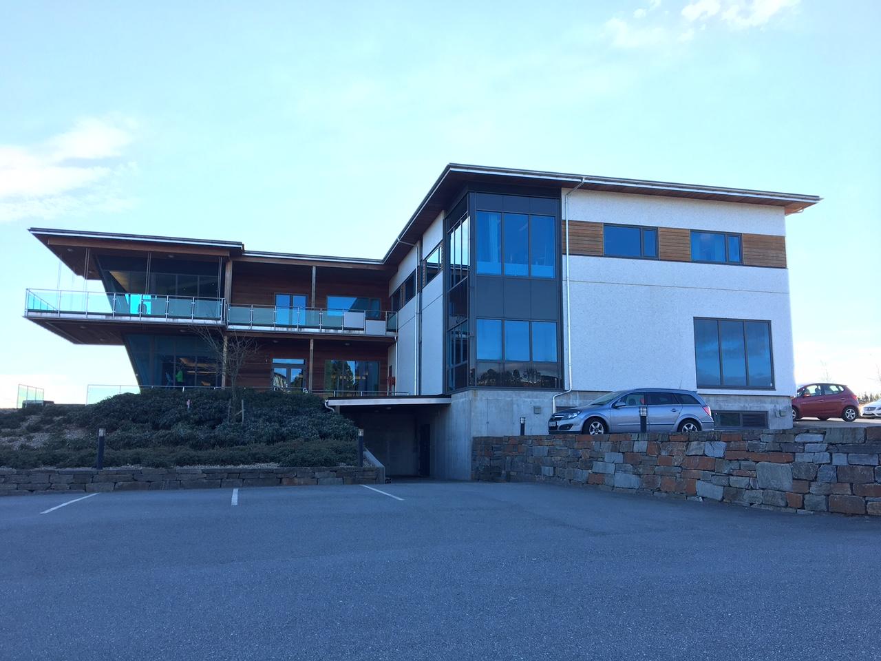 Moderne kontorbygg med store vinduer og terrasse, omgitt av parkeringsplasser og landskap.
