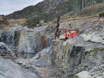 Gravemaskin arbeider i en steinbrudd med bratte skråninger og store steiner i omgivelsene.