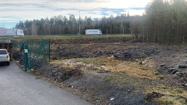 Byggeplass med grus og trær, biler i bakgrunnen, og arbeidere i oransje klær.