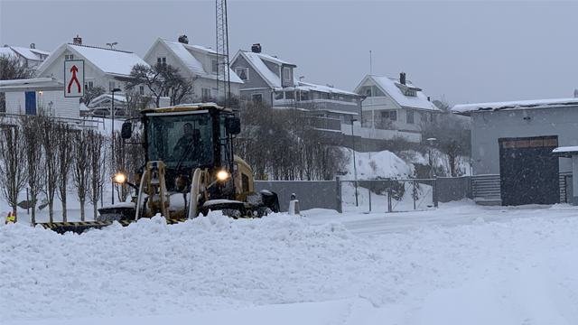 En snøplog rydder snø fra veien i et boligområde under snøvær. Hvit snø dekker bakken.