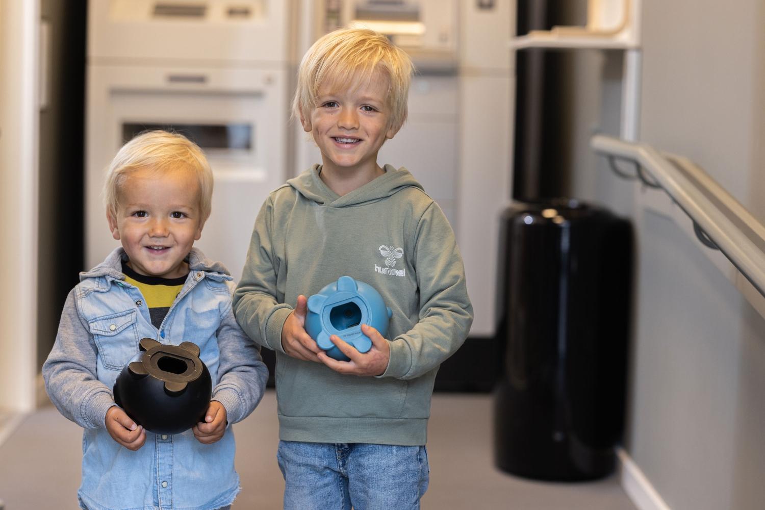 To barn smiler og holder fargerike leker i en moderne innendørs setting nær en minibank.