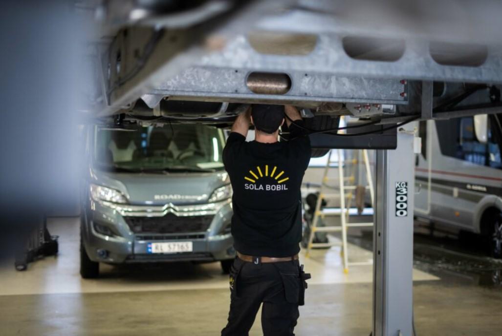 En mekaniker jobber under en bil i et verksted, med en Citroën i bakgrunnen.