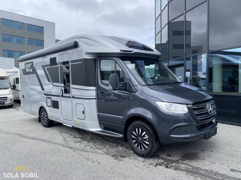 En moderne campingvogn parkert foran en bygning, med grå og sølvfarge og store vinduer.