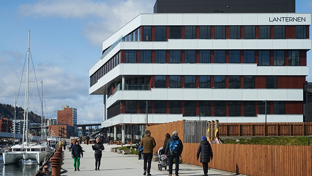 Moderne bygning med navnet "Lanternen" ved havnen, folk som går langs promenaden og båter i bakgrunnen.