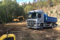 En grå lastebil med blå tilhenger står på en byggeplass, med en gravemaskin i bakgrunnen.