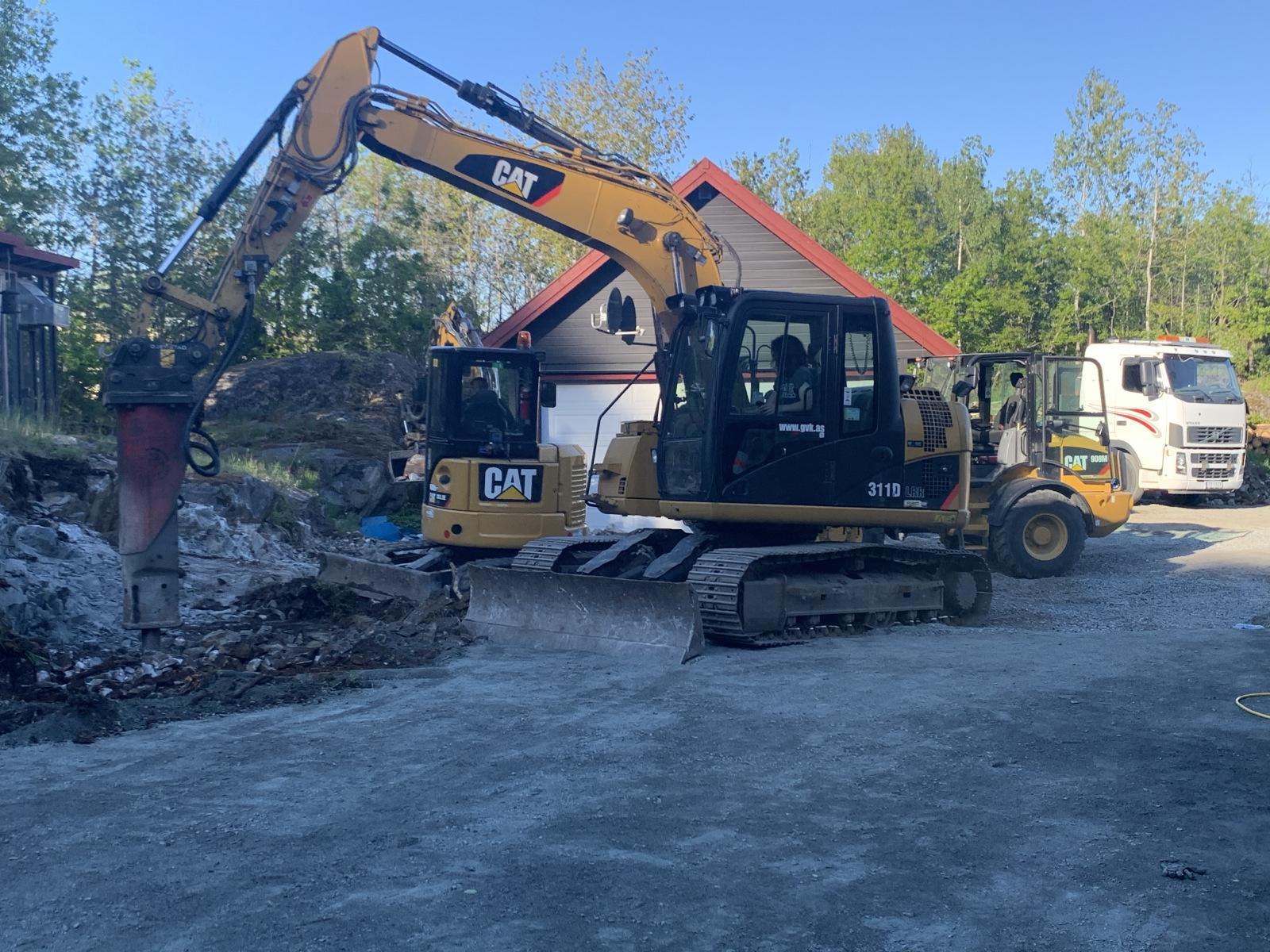 Gravemaskin og hjullaster i arbeid på byggeplass med hus i bakgrunnen og lastebil til høyre.