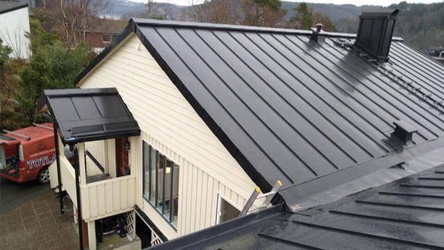 Tak på en moderne enebolig med svart metalltak og solcellepaneler, med en rød bil parkert i bakgrunnen.
