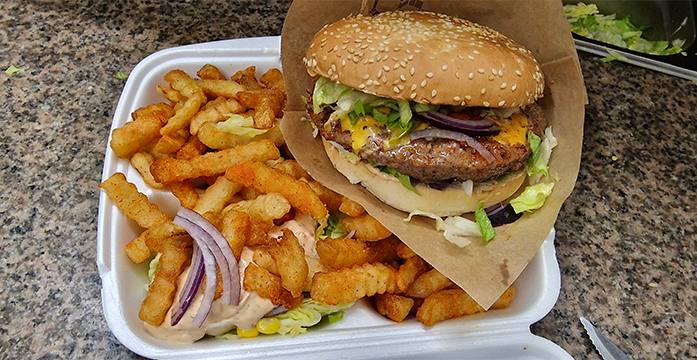 En saftig hamburger med sesamfrøbrød, salat, løk og pommes frites på en hvit plasttallerken.