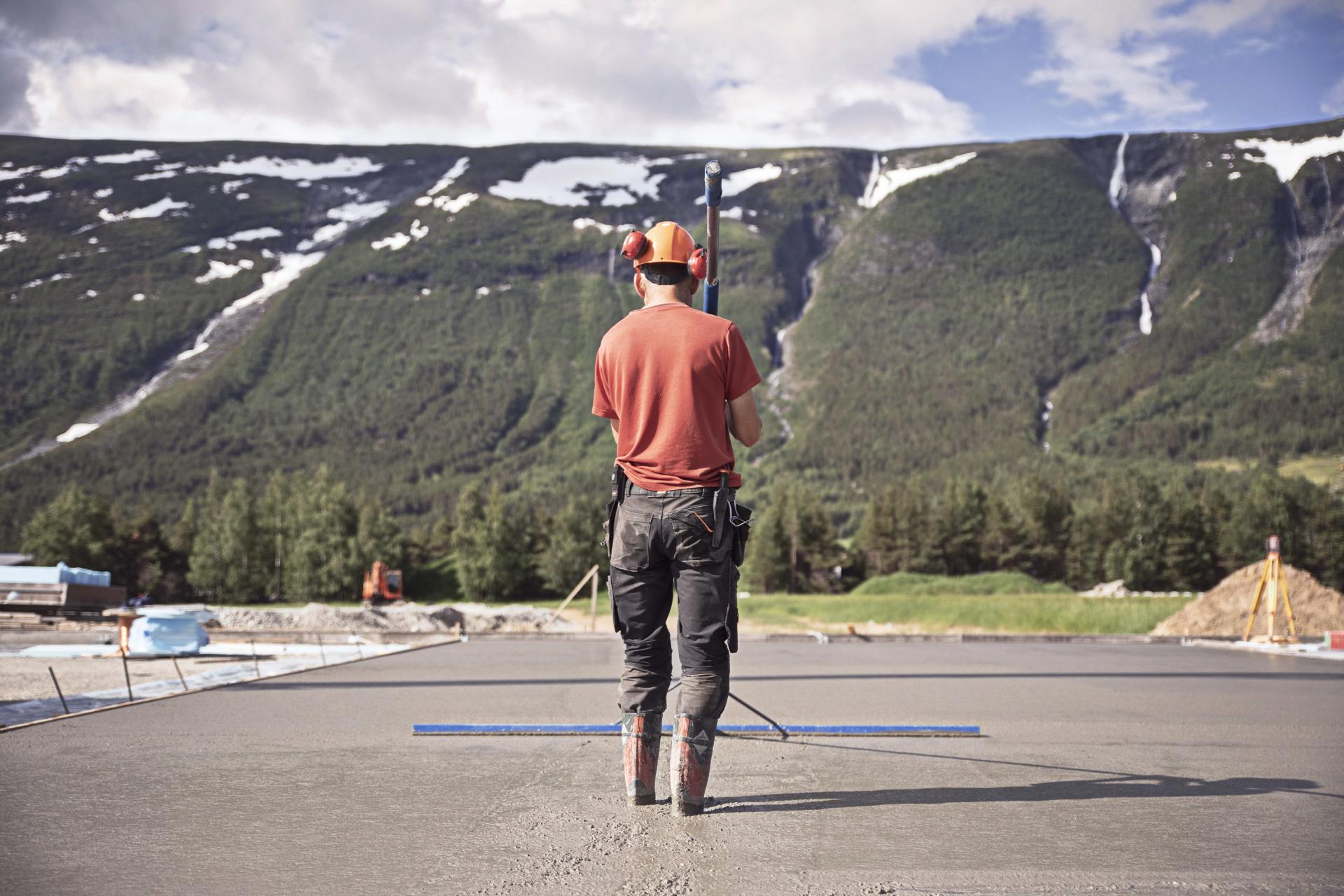 En mann i arbeid med en måler på en byggeplass, med fjell og fossefall i bakgrunnen.