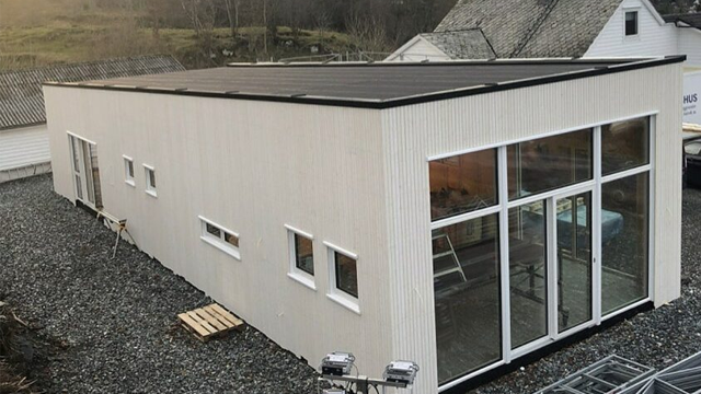 Moderne enebolig med store vinduer og solcellepaneler på taket, omringet av grus og natur.