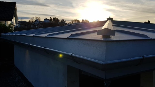 Tak med metallbelegg og solnedgang i bakgrunnen, med en klar himmel og bygninger i det fjerne.