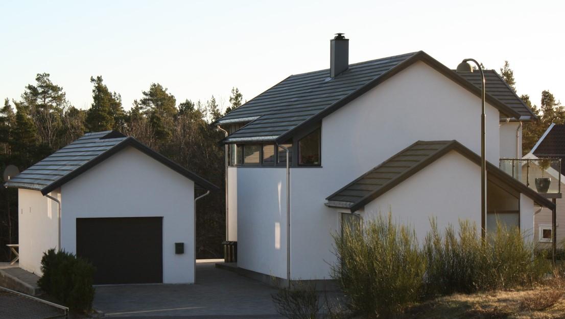 Moderne hus med garasje og hage, omgitt av trær i sollys.