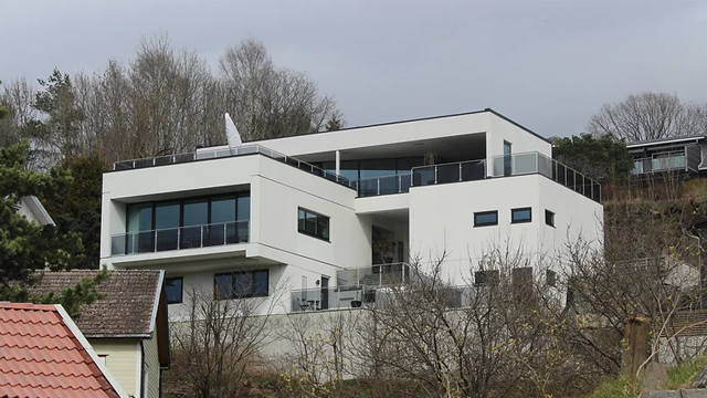 Moderne, hvit enebolig med store vinduer og terrasse, beliggende på en skråning med trær i bakgrunnen.