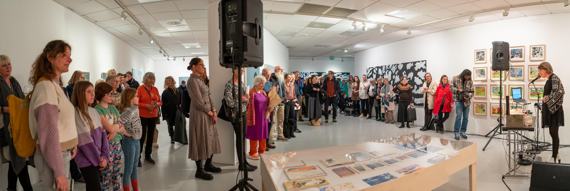 En stor gruppe mennesker står i et kunstgalleri under en utstilling, med kunstverk på veggene og en taler foran.