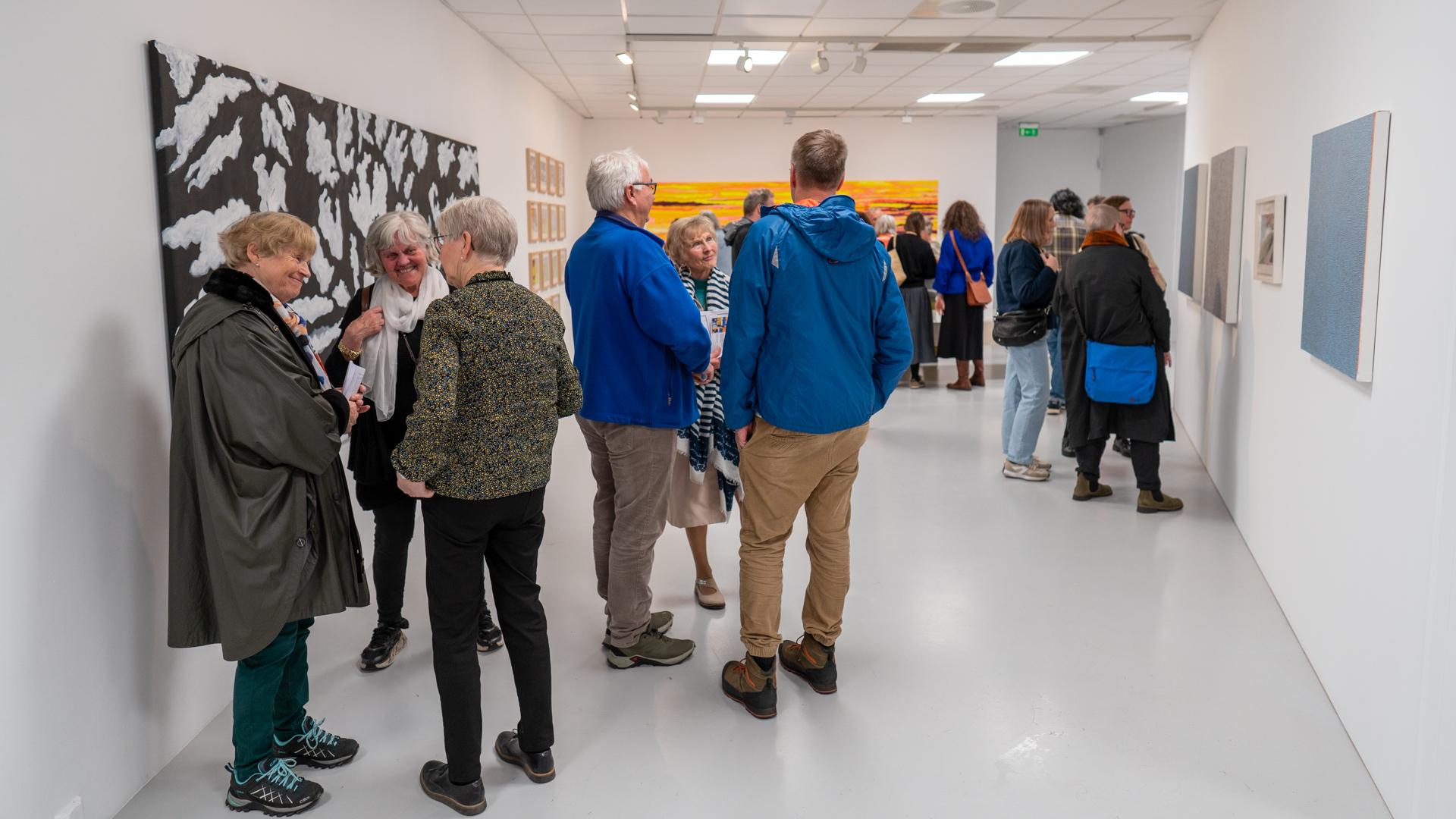 Besøkende i et kunstgalleri, med flere kunstverk på veggene og folk som samtaler i rommet.