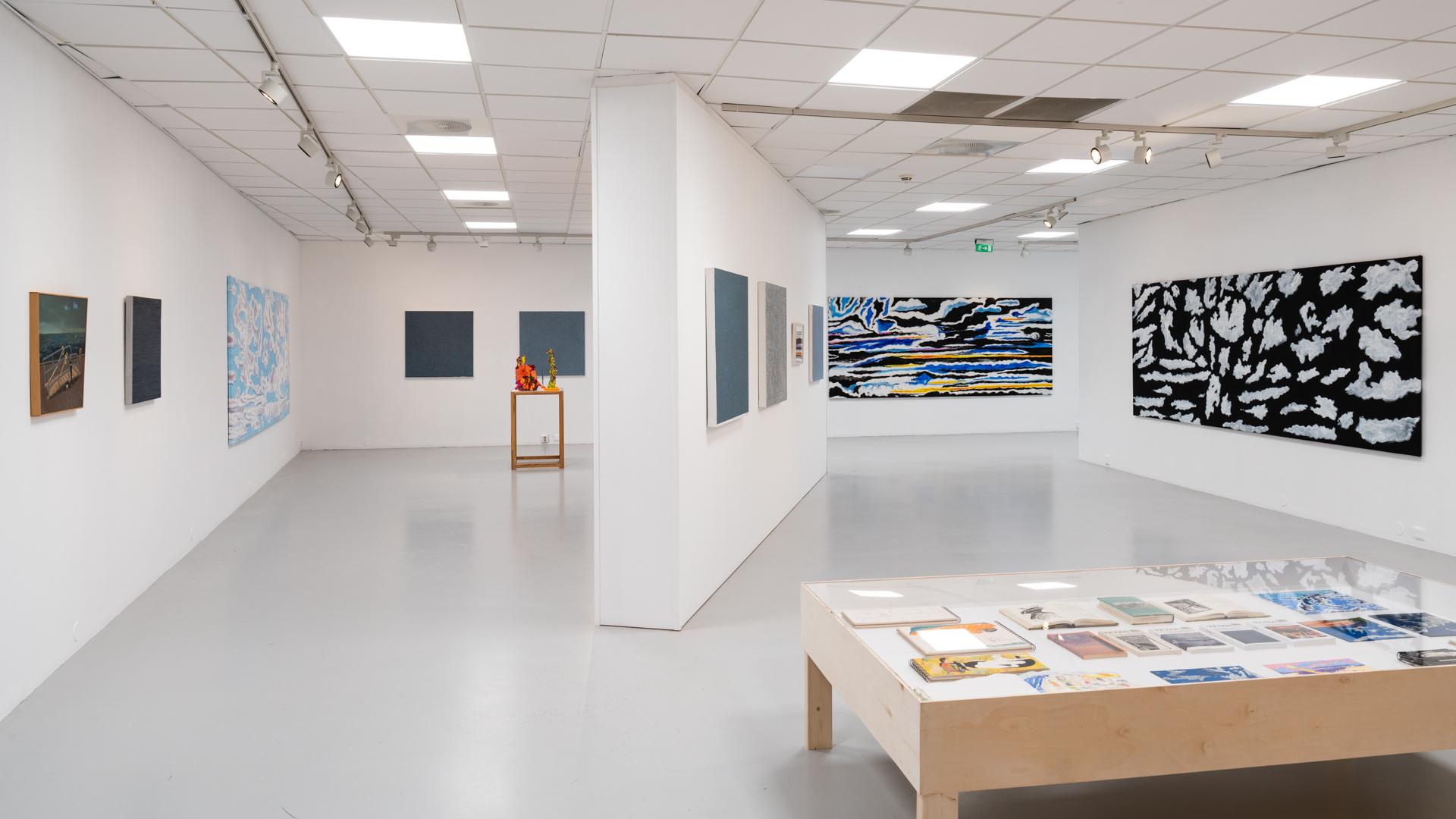 Kunstgalleri med malerier på veggene og et bord med kunstverk i midten av rommet.