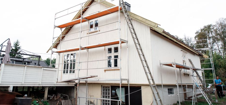 Hus med stillas rundt, under oppussing, med vinduer og tak i lys farge.