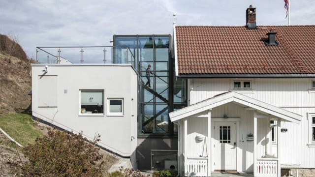 Moderne hus med glassfasade og tradisjonelt hvitt hus ved siden av, beliggende i naturskjønt område.