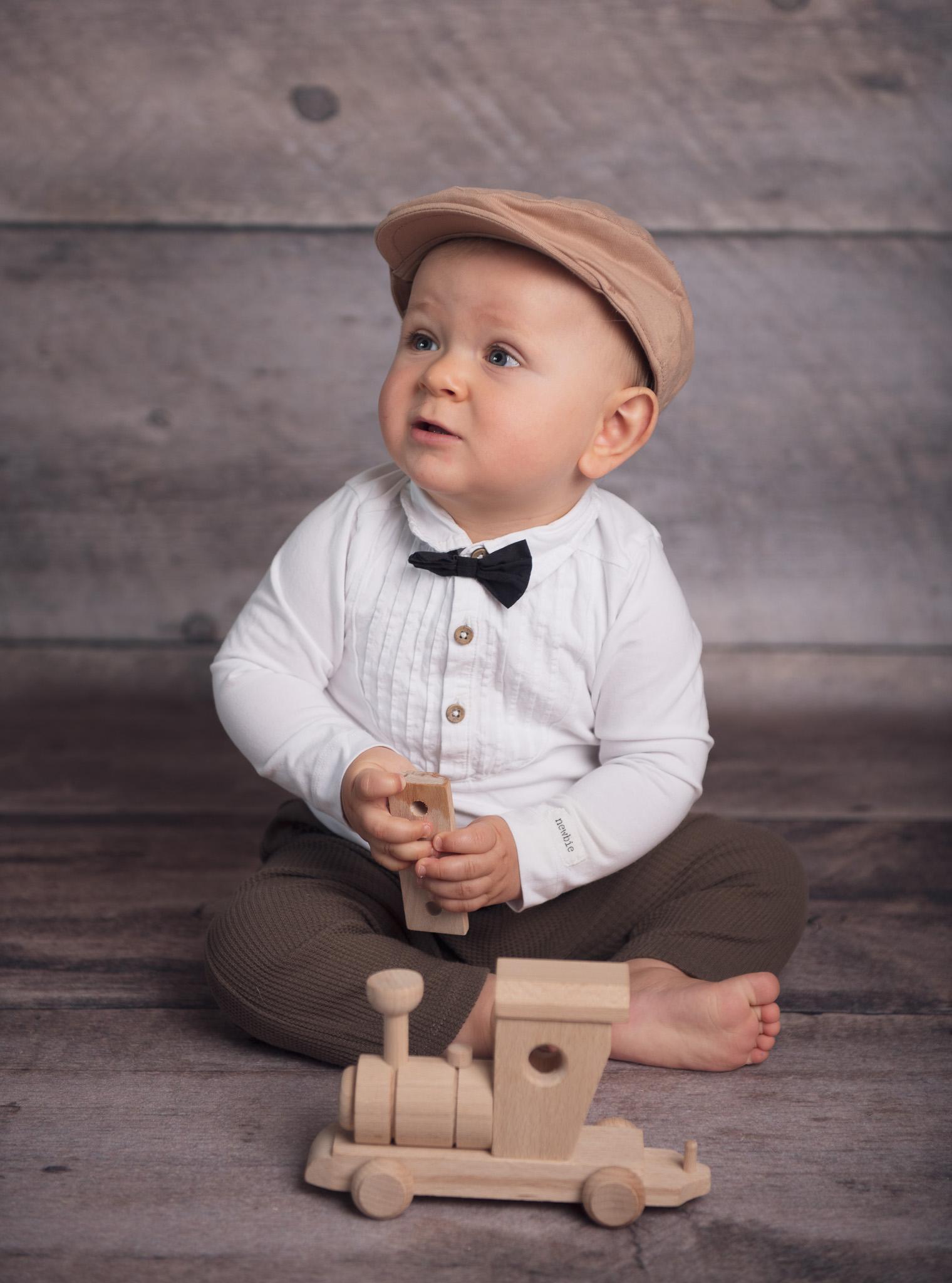 En liten gutt sitter på gulvet med en treleke i form av et tog, iført en hvit skjorte og en beige caps.