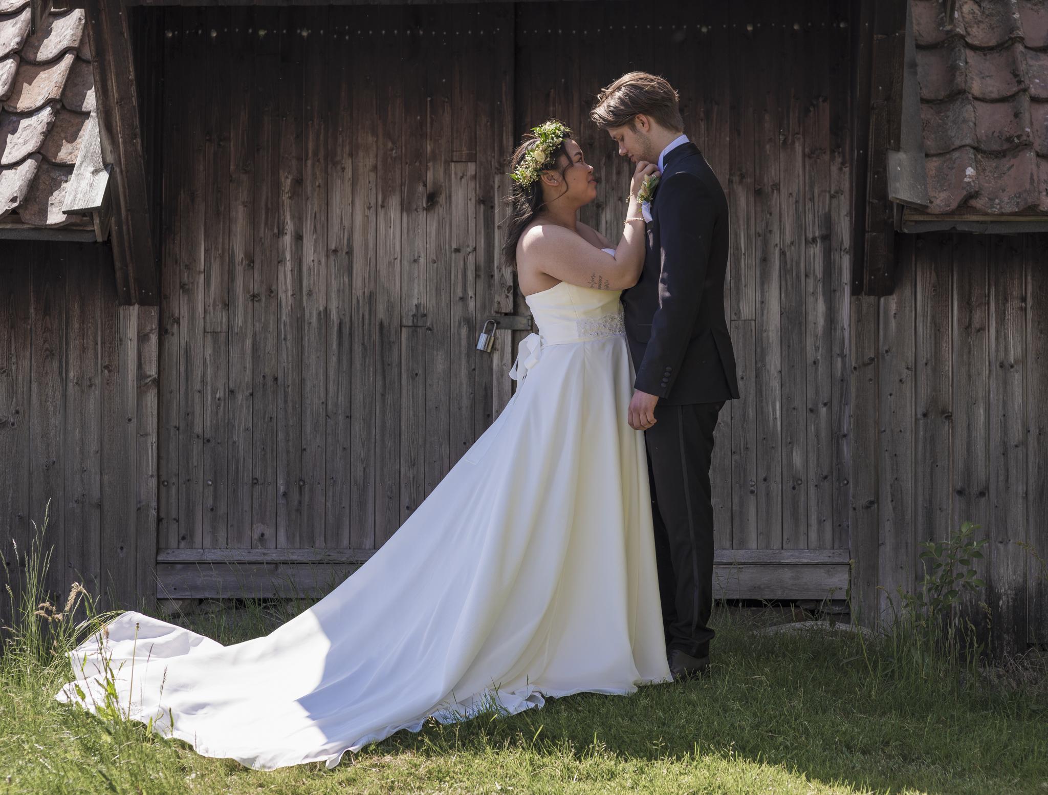 Brudeparet står foran en gammel trevegg, bruden i hvit kjole med blomsterkrans, brudgommen i mørk dress.