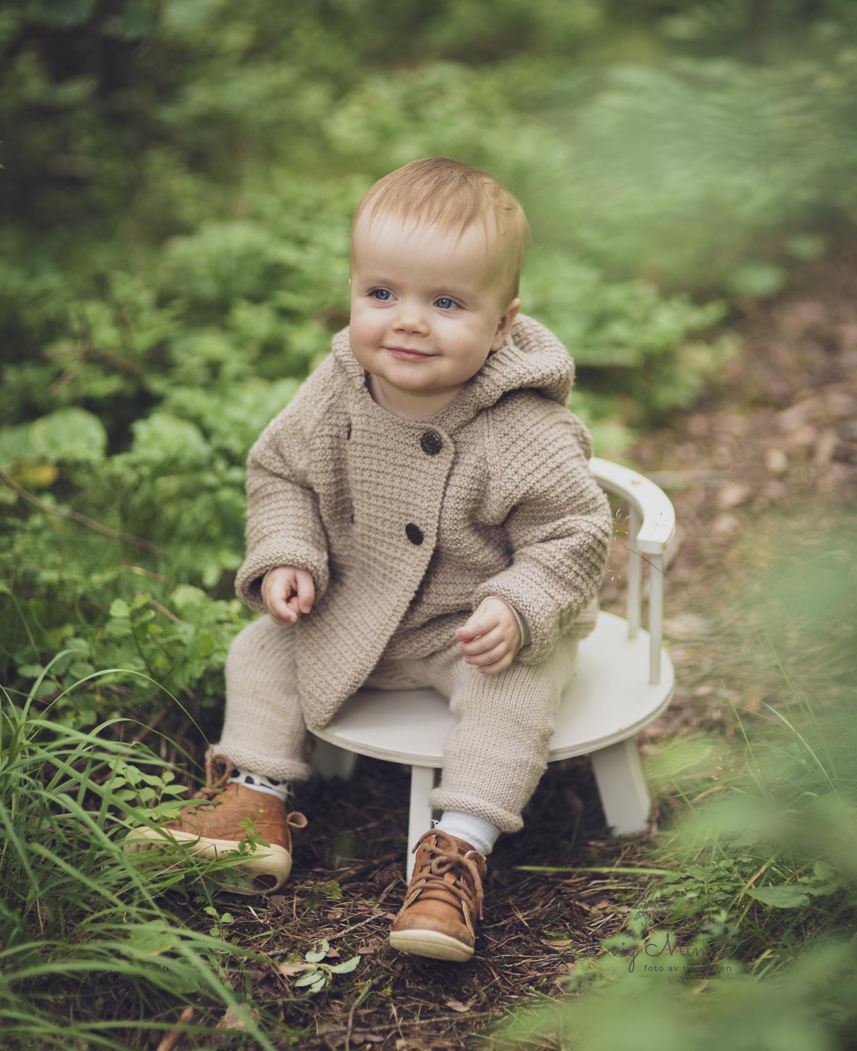 Liten gutt sitter på en hvit stol i skogen, iført en beige genser og brune sko, omgitt av grønt gress.