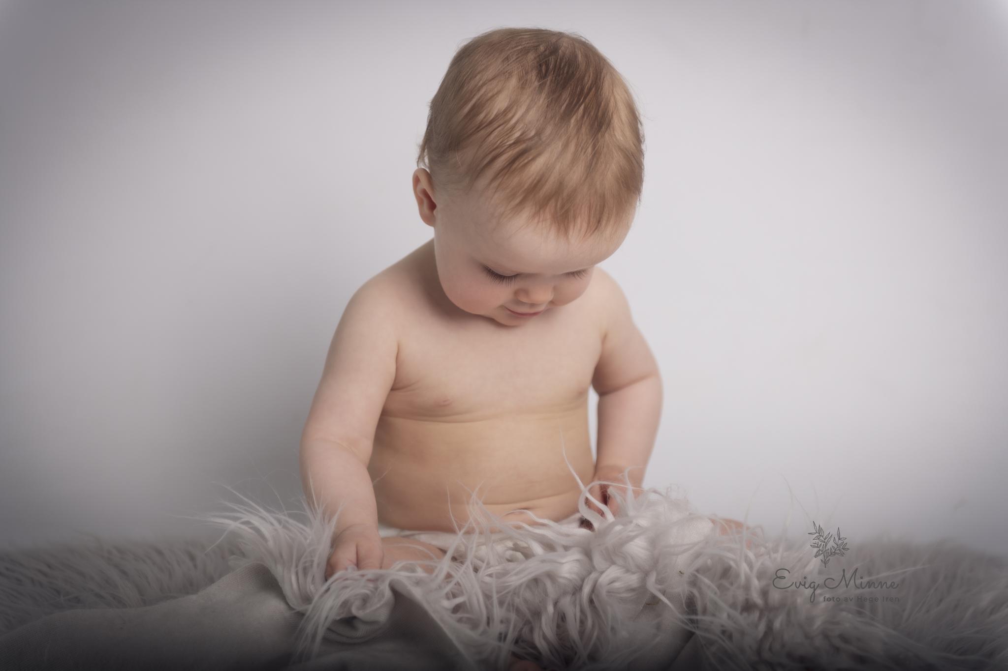 Liten baby sitter på en myk, grå pels, ser ned på stoffet med nysgjerrige øyne.