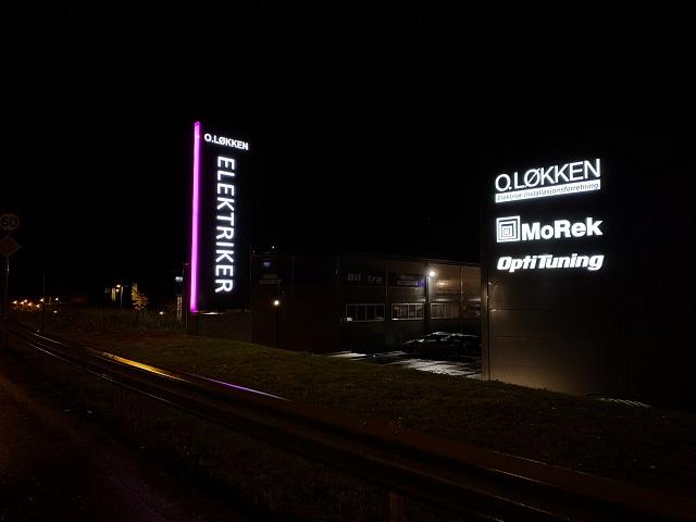 Lysende skilt for QLøkken Elektrikere og MoReK OptiRunning ved en vei om natten.