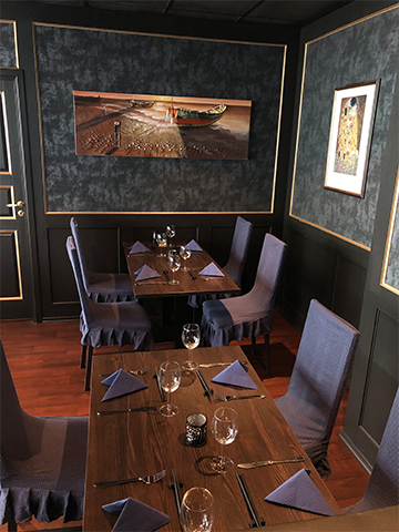En koselig restaurant med mørke vegger, bord dekket med blå servietter og glass, og et maleri på veggen.