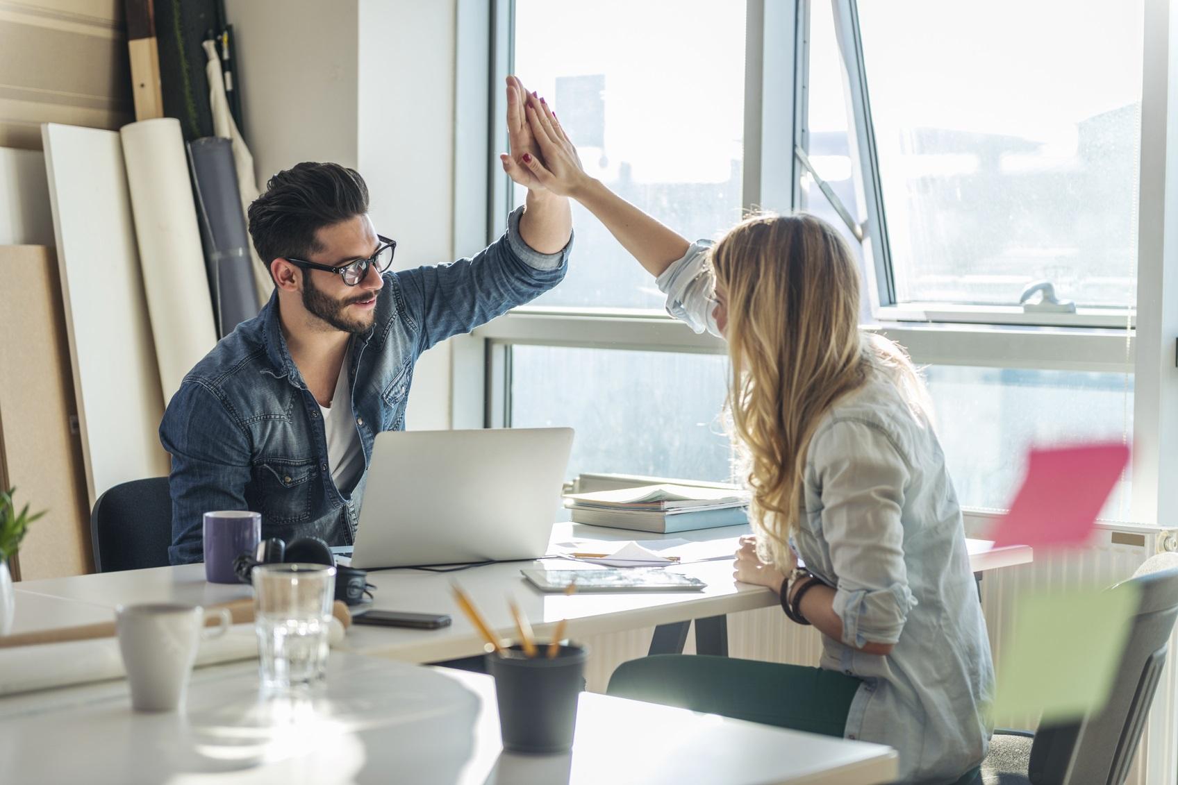 To personer i et kontor, en mann og en kvinne, gir hverandre high five over et skrivebord med en laptop.