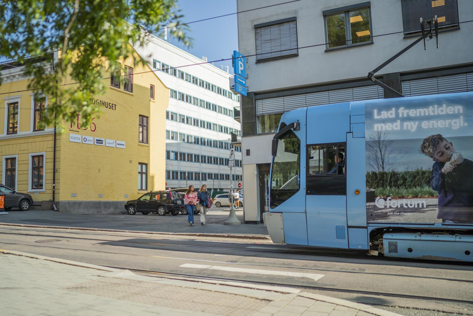 Trikk passerer en gate med bygninger og parkerte biler, med en reklameplakat på siden av trikken.