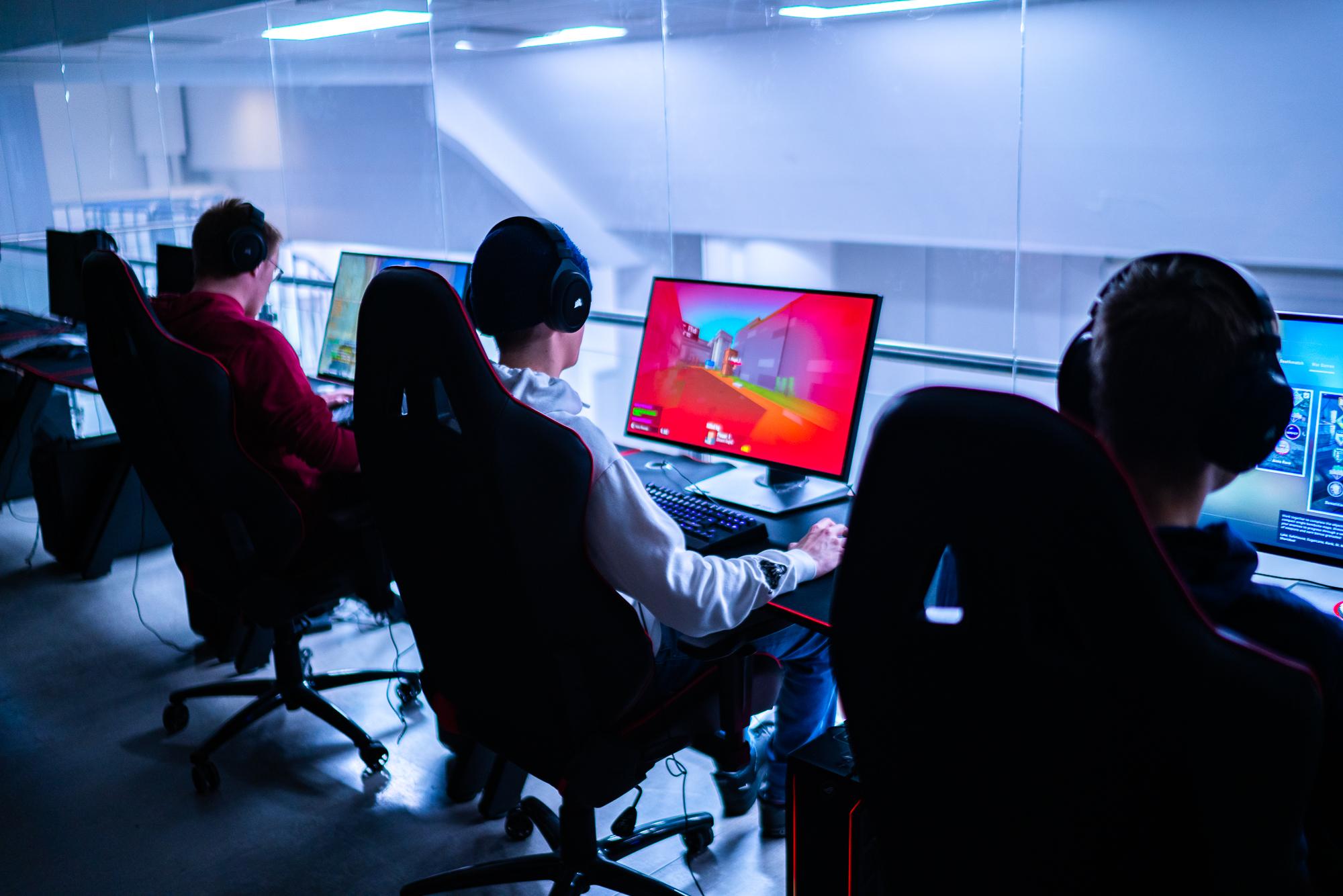 Tre personer sitter i gamingstoler og spiller videospill på datamaskiner i et moderne rom.