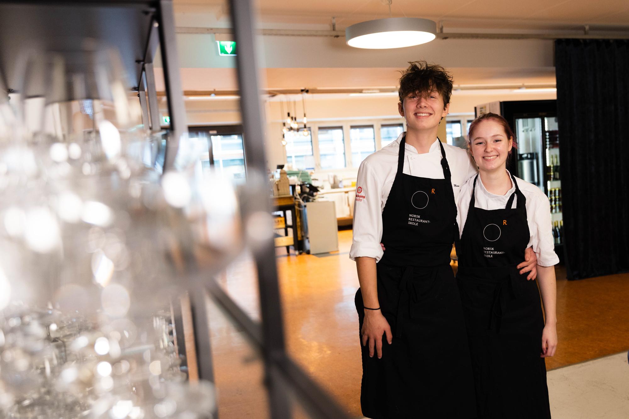 To kokker i hvite skjorter og svarte forkle står smilende i en restaurant med glass i bakgrunnen.