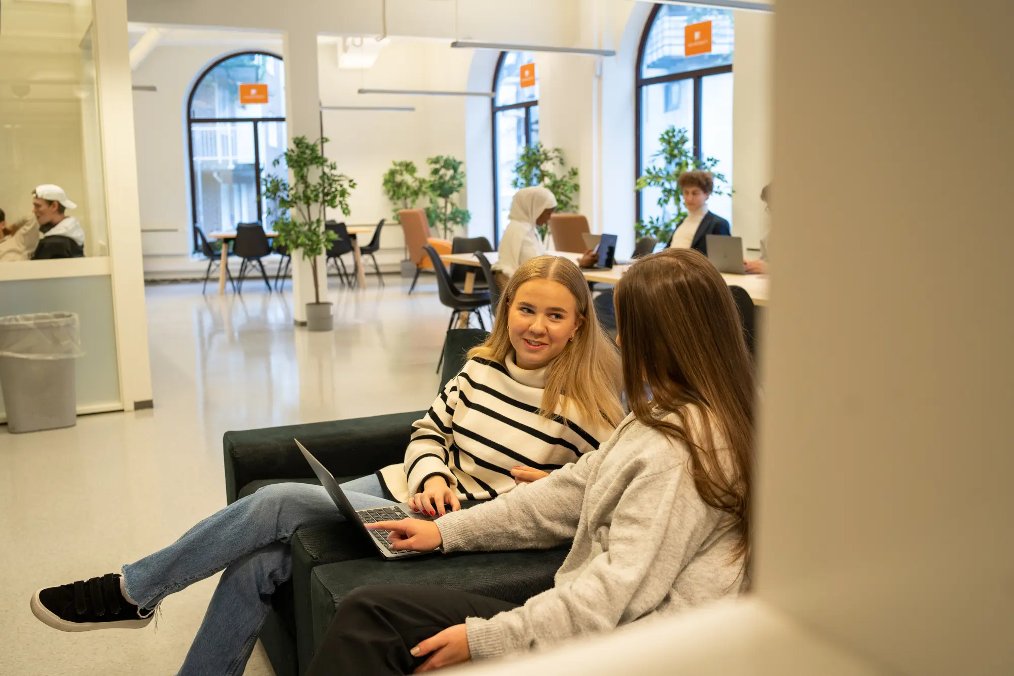 To jenter sitter på en sofa og jobber på en laptop i et moderne kontormiljø med planter rundt.