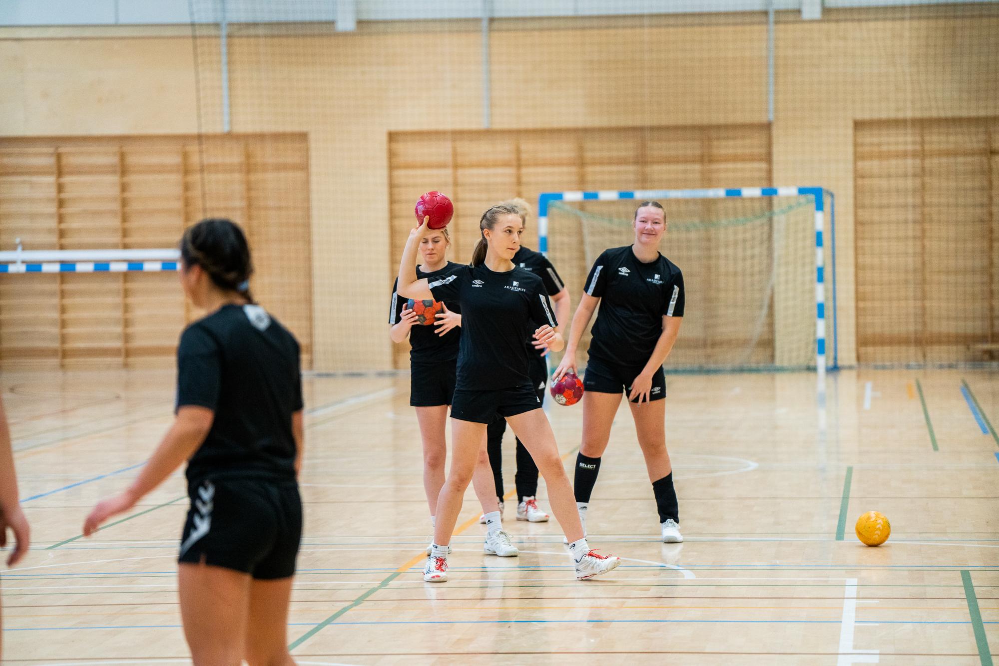 Fire jenter trener håndball i en gymsal, iført svarte drakter og holder på med balløvelser.
