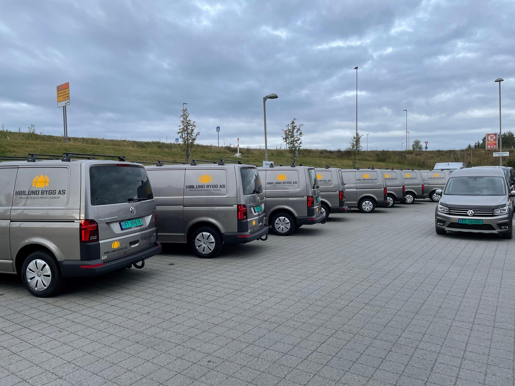 Flere grå varebiler parkert på en asfaltplass med trær i bakgrunnen.