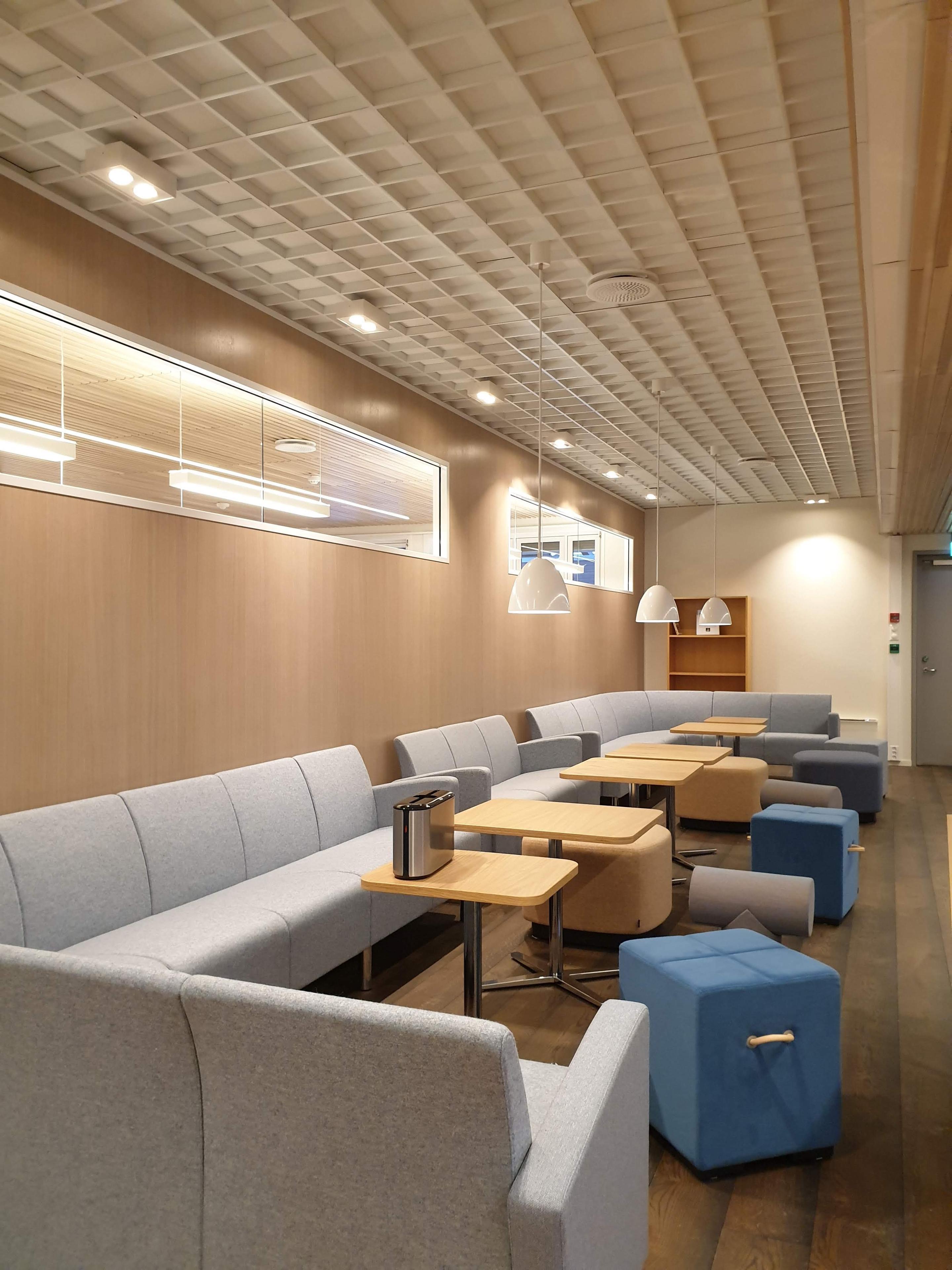 Moderne lounge med grå sofaer, trebord og blå puffer, belyst av taklamper i et stilrent rom.