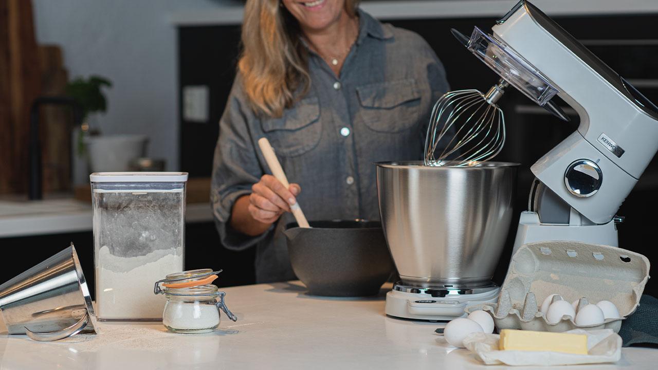 En kvinne som baker med en kjøkkenmaskin, mel, smør og egg på kjøkkenbenken.