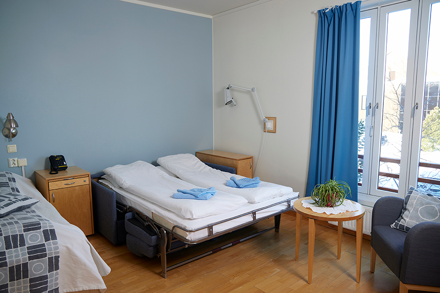Lyst rom med to enkeltsenger, blå gardiner, en stol og et lite bord med planter.