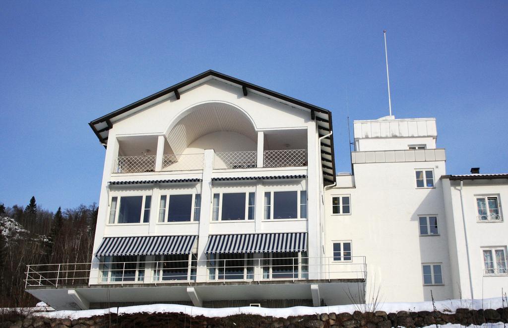 Moderne hvitt hus med balkonger og store vinduer, omgitt av snø og trær under en klar blå himmel.