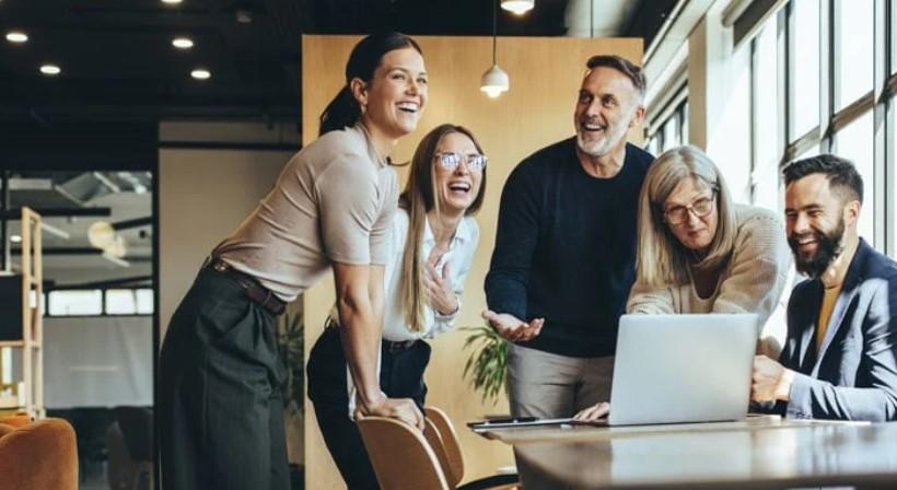 En gruppe glade kolleger samles rundt en laptop i et moderne kontormiljø.