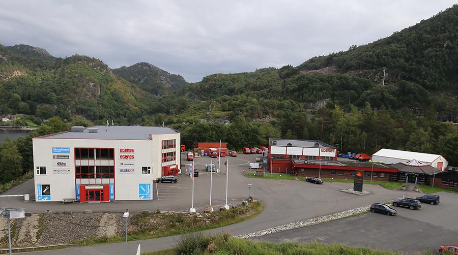 Bilde av et forretningsområde med flere bygninger, parkerte biler og grønne fjell i bakgrunnen.