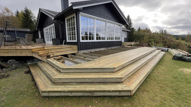 Terrasse med flere nivåer i tre, ved siden av en moderne hytte med store vinduer og grønt landskap i bakgrunnen.