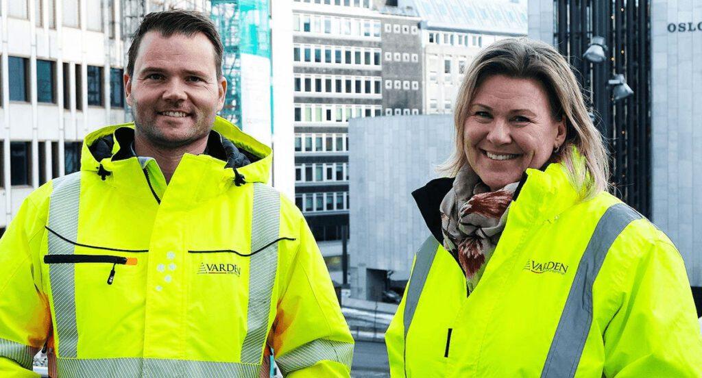 To personer i gule arbeidsjakker står ute i byen, med moderne bygninger i bakgrunnen.