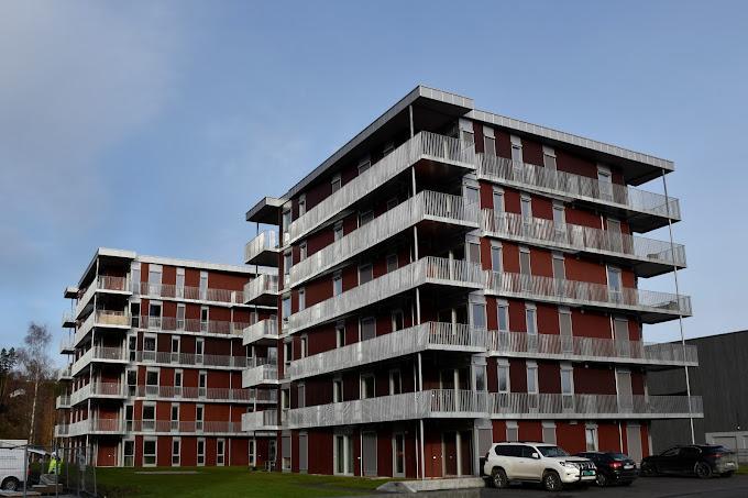 Moderne leilighetsbygg med flere etasjer og balkonger, omgitt av grønt område og blå himmel.