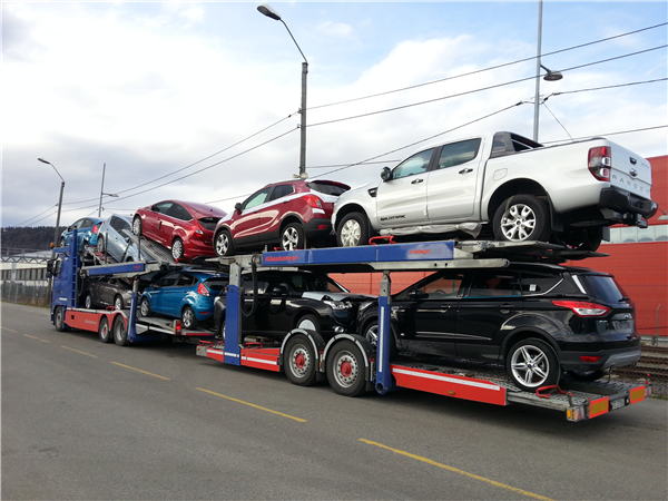 En biltransport med flere biler stablet på to nivåer, inkludert SUV-er og en pickup, parkert langs veien.