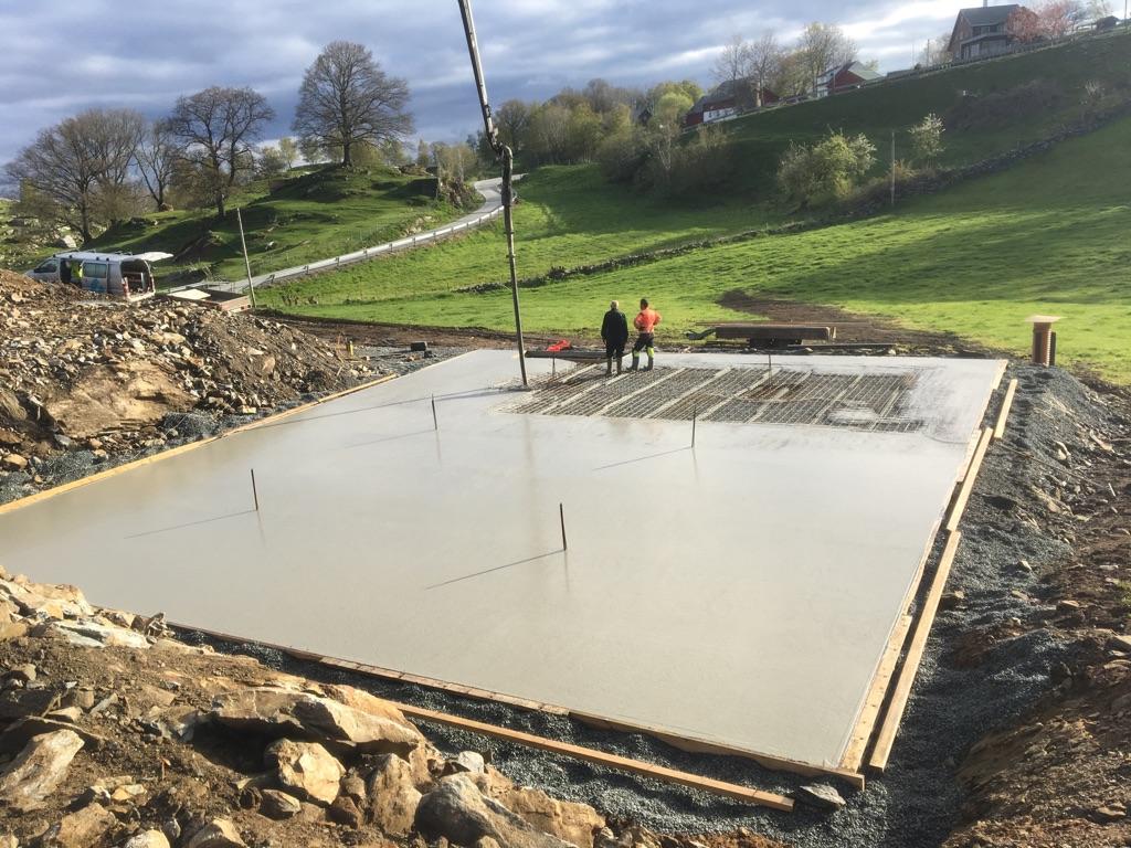 Betongplaten legges på byggeplassen med arbeidere og maskiner i bakgrunnen, omgitt av grønt landskap.